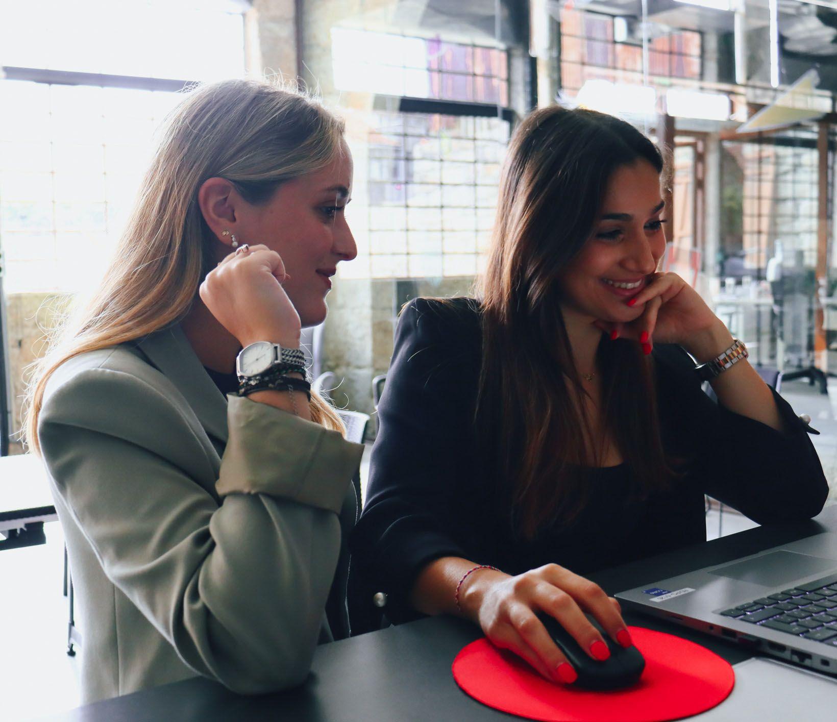 two young women working side by side at the computer with big smiles. On the left side, a blonde woman dressed in shades of green and on the right side of the image, a brunette woman dressed in black. In the background, we can see the white wall of the office decorated with industrial details.