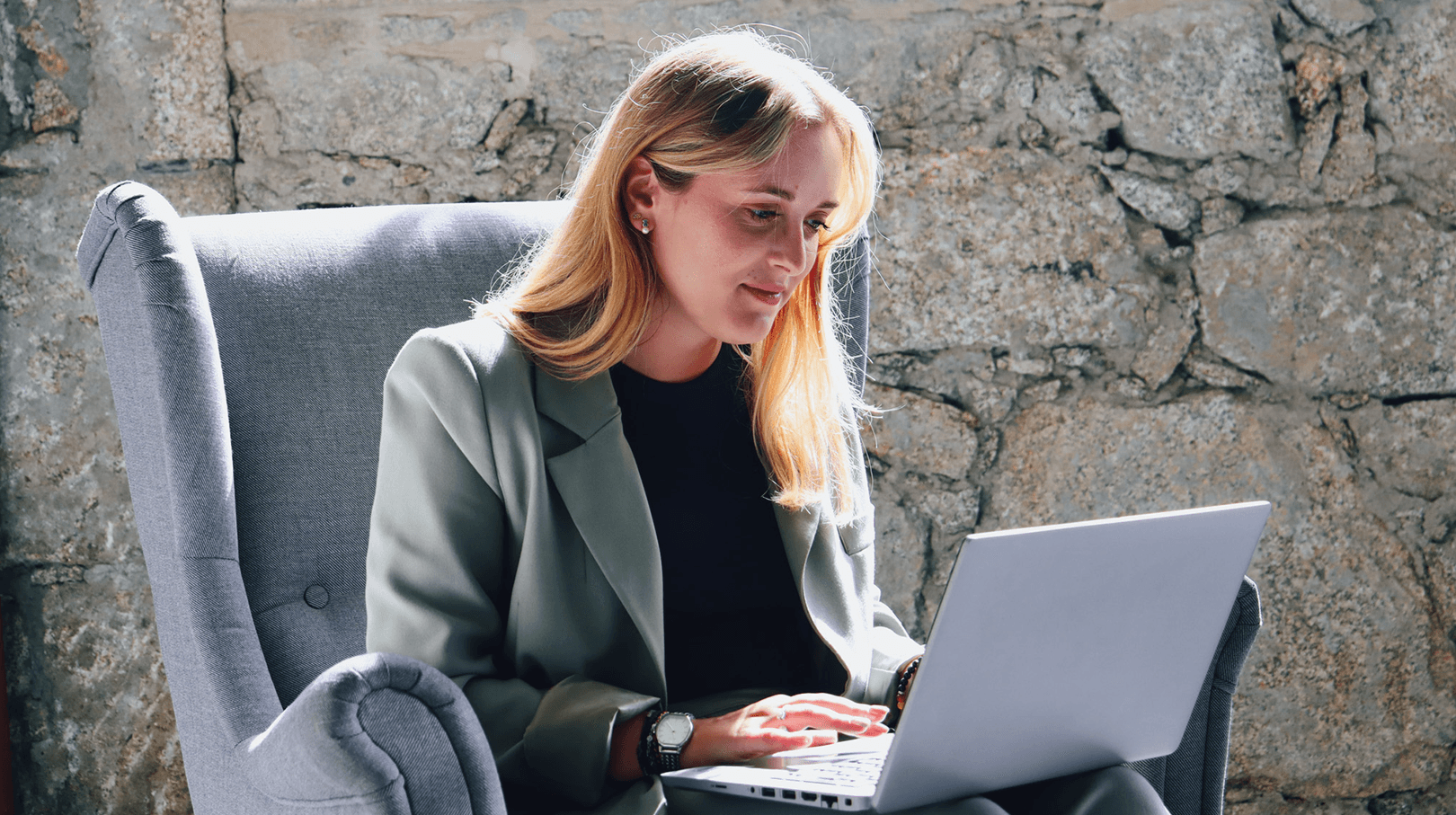 young woman working at her laptop sitting on a comfy chair. The backgound is a wall of stone.