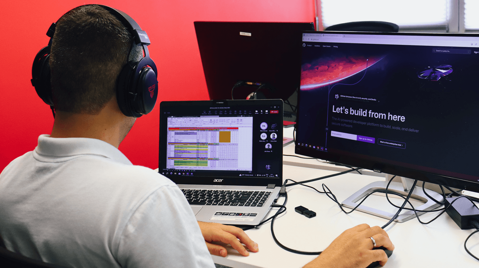 young man with his back turned, looking at his laptop and with an image on a second, larger computer screen. In the background we can see the office decoration in tones of red and white.