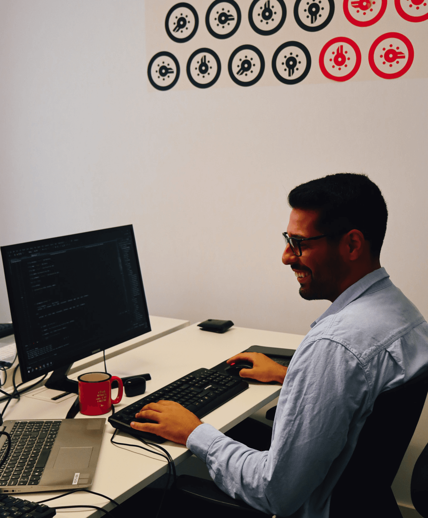 young man smiling while working on programming on his laptop and computer screen. In the background we can see the ton's office decoration