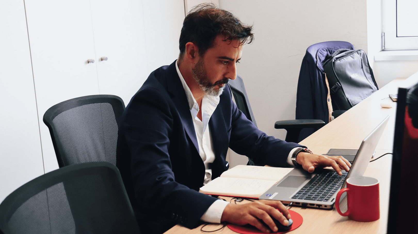 man focused on his work on the computer, looking concentrated and dressed in a blue suit and white shirt, with a watch on his left wrist. His desk at work has a red mug on top. In the background, we can see the office with two other working chairs.