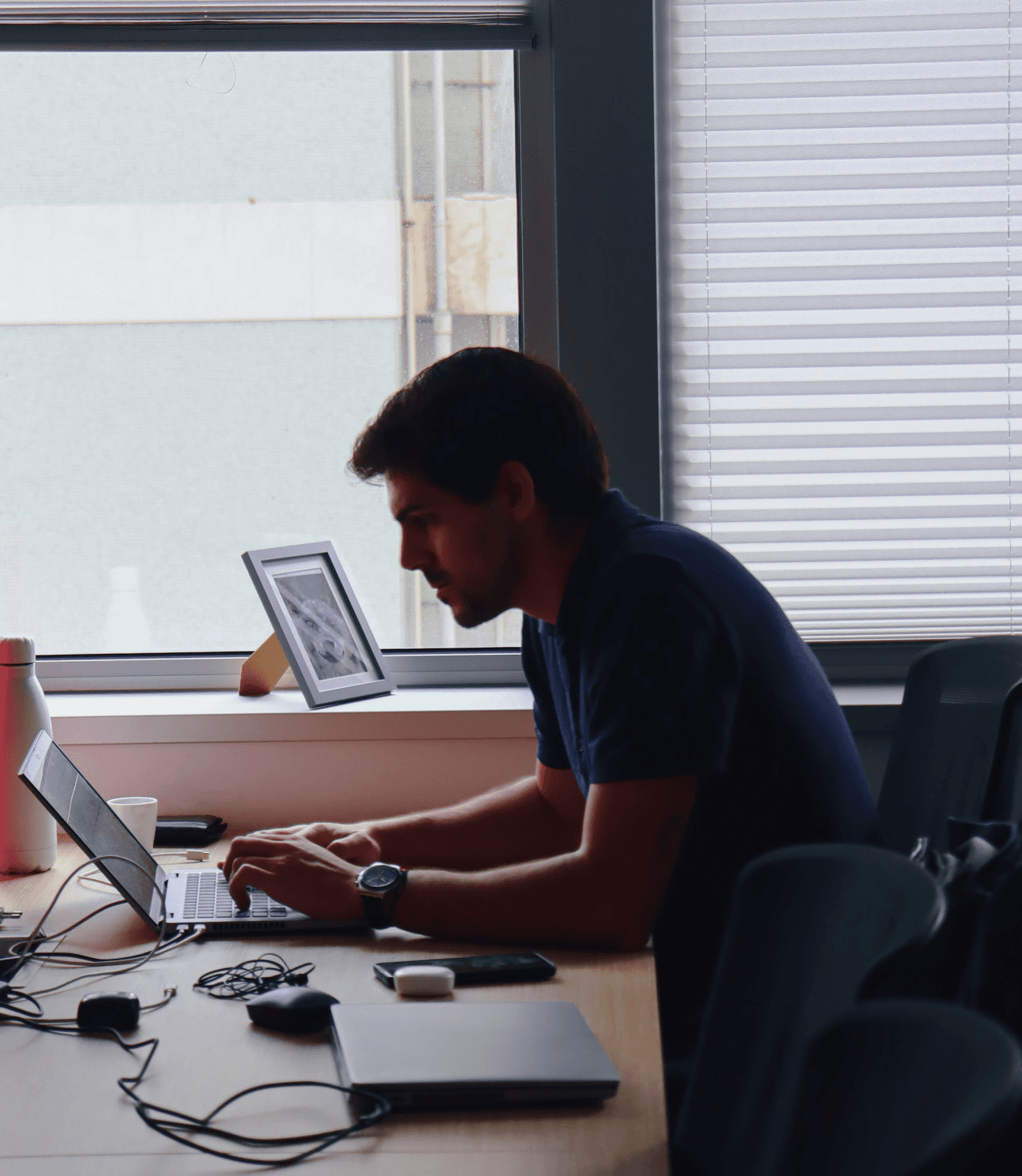 young man focused and working on the computer, dressed in a blue shirt. In the background, a window to outdoors.