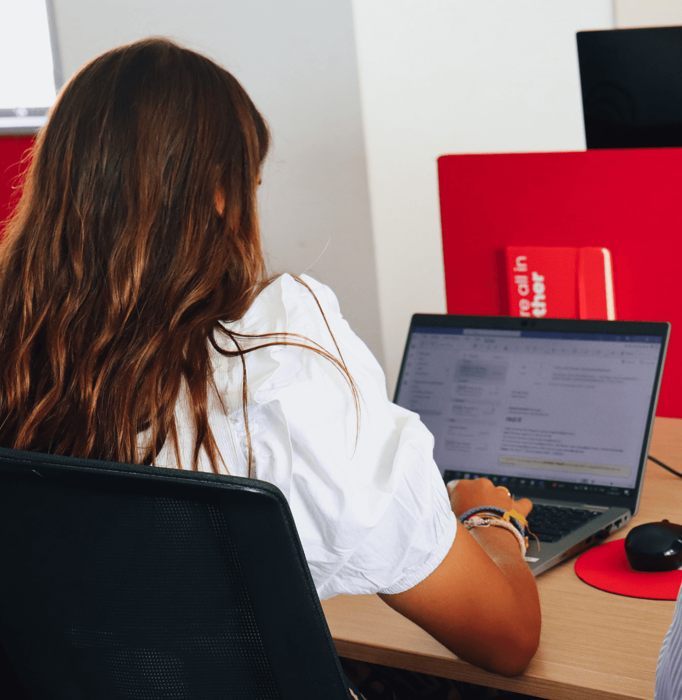 young brunette woman from behind, working on the computer. In the background, the work table with red details