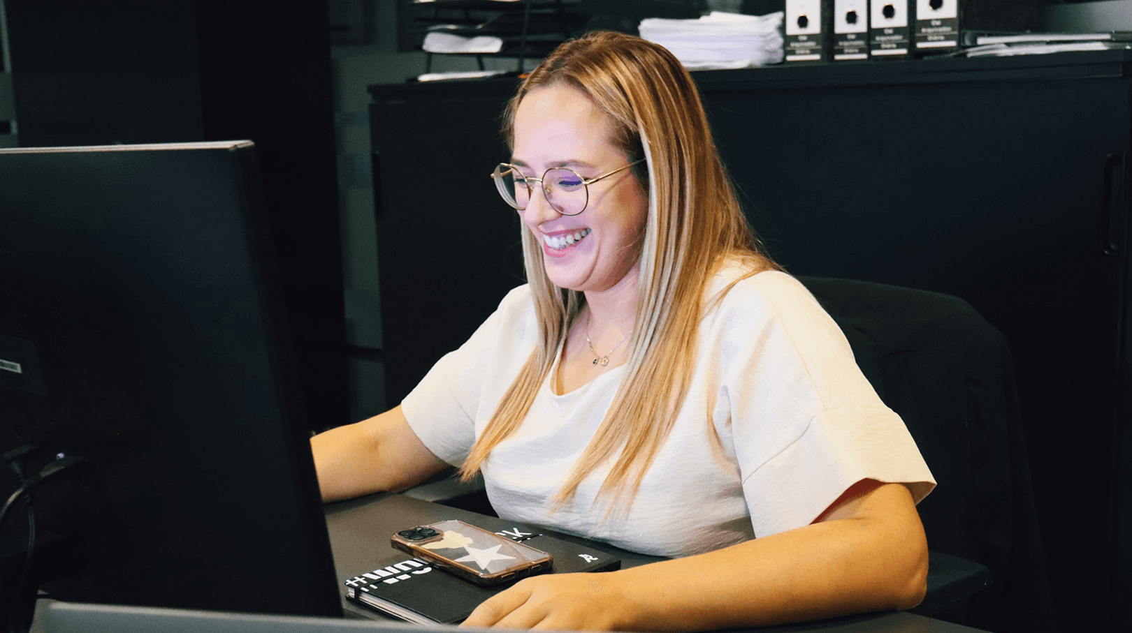 young blonde woman smiling while working at the computer. Dressed in shades of white. In the background we can see details of the office, all in shades of black.
