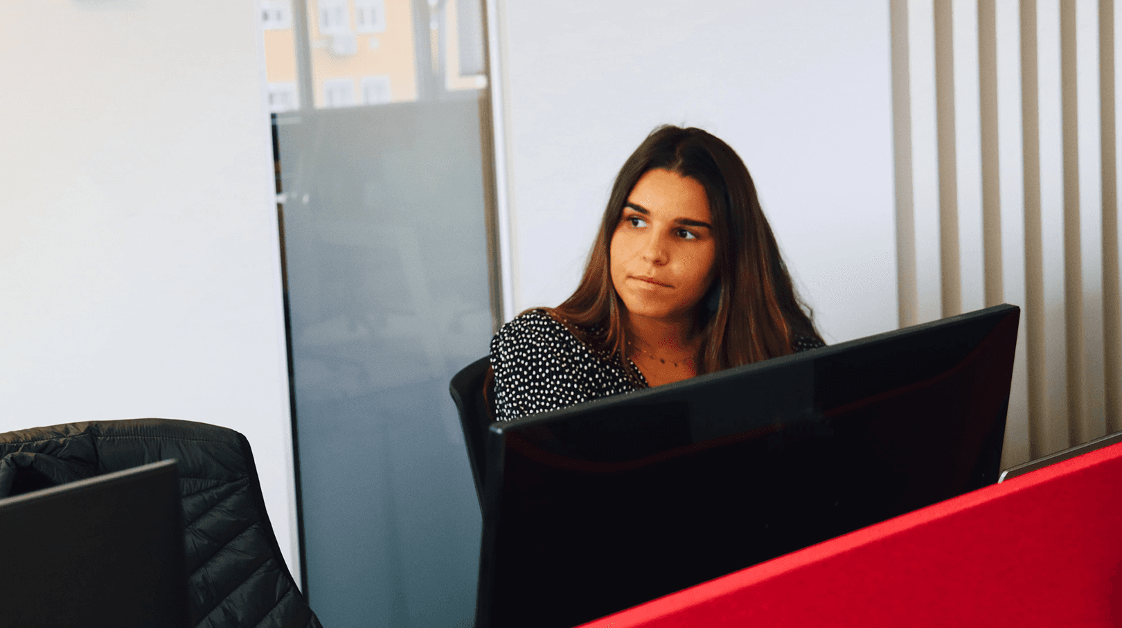 picture of a young woman with a computer screen in front of her, looking to the right, with a focused and interested look. The woman is brunette and is dressed in black. In the background we can see the office in white and red tones.